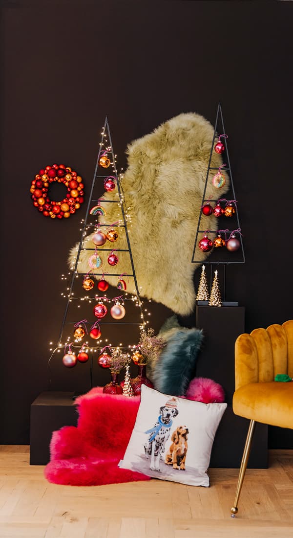 Weihnachtsdeko mit bunten Kugeln an metallischen Baumgestellen, rotem Kranz, Fell und Kissen mit Hundemotiv, gelber Sessel