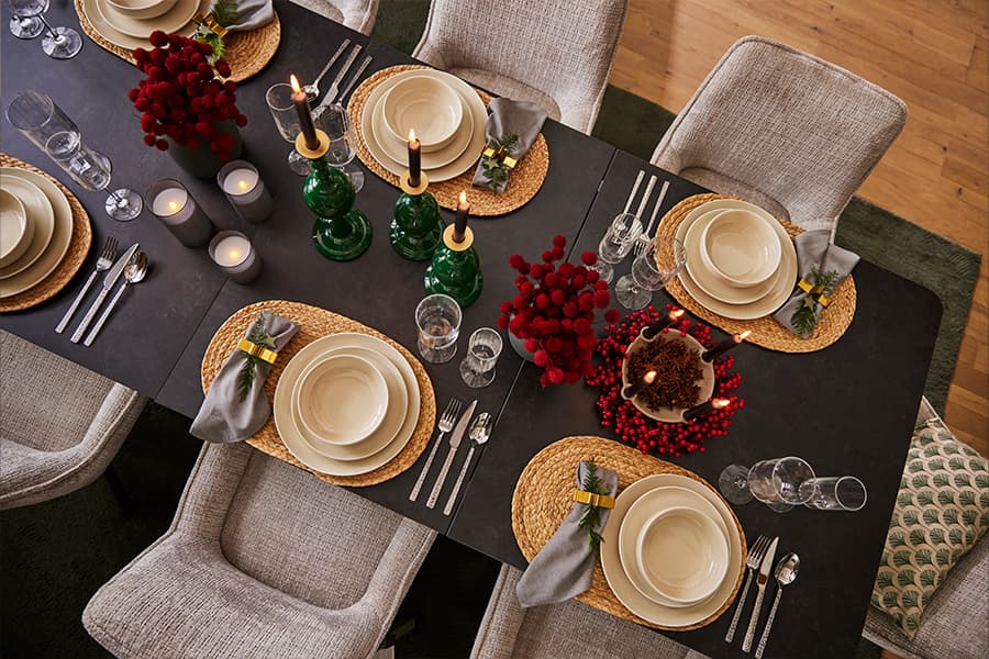 Festlich gedeckter Tisch mit Tellern, Besteck und Gläsern, roten Deko-Beeren, Kerzen, grünen Kerzenhaltern und grauen Servietten.