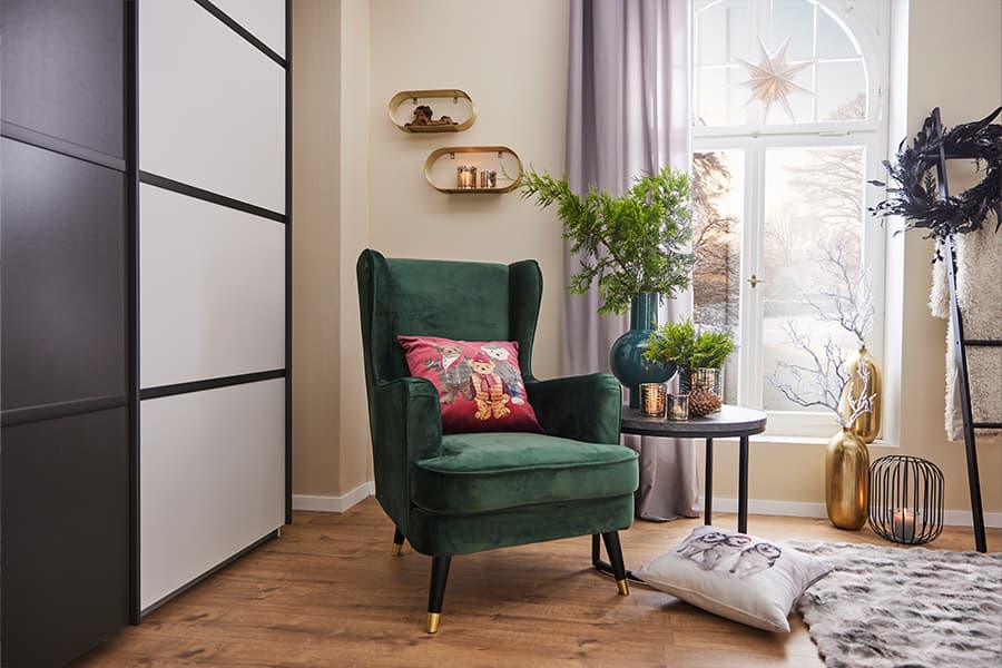 Gemütliches Wohnzimmer mit grünem Sessel, rundem Tisch, Pflanzen und Dekorationen. Kissen mit weihnachtlichem Motiv auf dem Sessel.