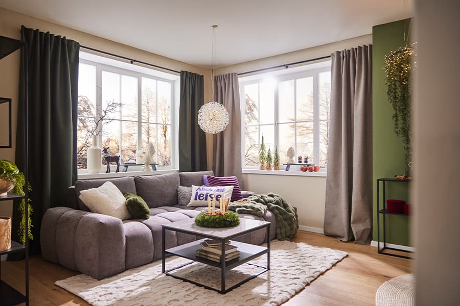 Wohnzimmer mit grauem Sofa, Kissen mit Aufschrift "Alles auf leise", Fenster mit Vorhängen, Couchtisch und Weihnachtsdekoration