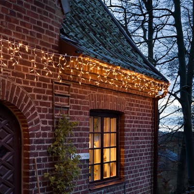 Gemütliches Backsteinhaus bei Dämmerung, Fenster beleuchtet, Lichterkette hängt am Dach, kletternde Pflanze an der Wand