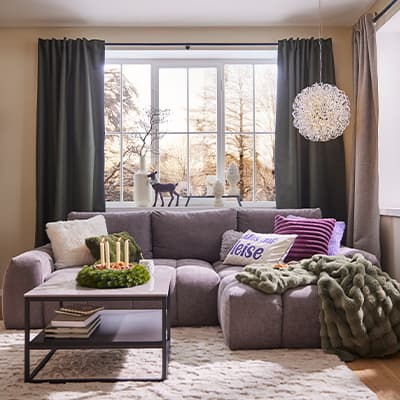 Gemütliches Wohnzimmer mit grauem Sofa, Kissen, Decke, Couchtisch und Adventskranz. Große Fenster mit grünen Vorhängen.