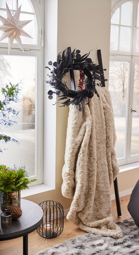 Schwarzer Kranz mit roten Akzenten hängt an einer Leiter, daneben eine flauschige Decke. Fenster und Weihnachtsdeko im Hintergrund.