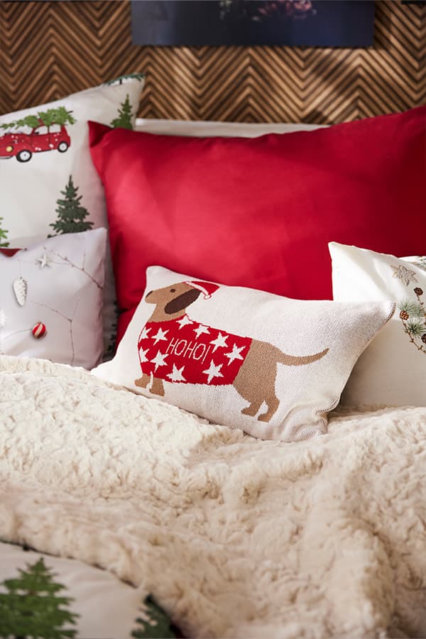 Kissen mit Hund und "HOHO!" Schriftzug, umgeben von roten, weißen und weihnachtlichen Kissen auf einem Bett mit weißer Decke