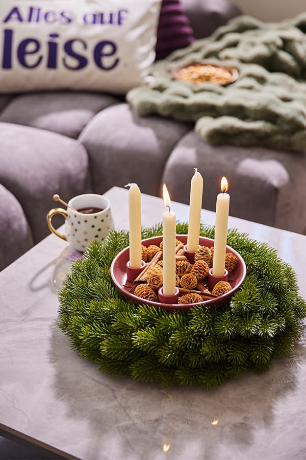 Alles auf leise – Sofa mit Kissen, Adventskranz mit vier weißen Kerzen, eine Tasse Tee auf dem Tisch, gemütliche Atmosphäre
