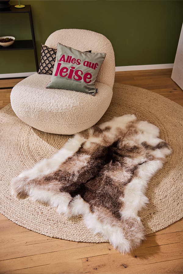 Alles auf leise – Graues Kissen mit rotem Text auf einem beigen Sessel neben einem gemusterten Kissen und einem Teppich mit Felloptik