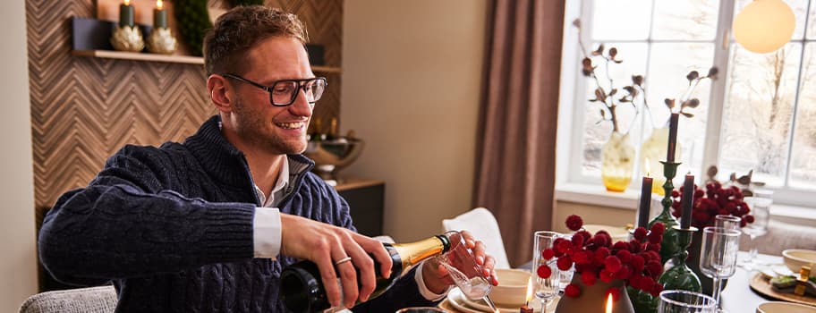 Mann in blauem Pullover öffnet eine Flasche Sekt am festlich gedeckten Tisch mit Kerzen und roten Blumen.