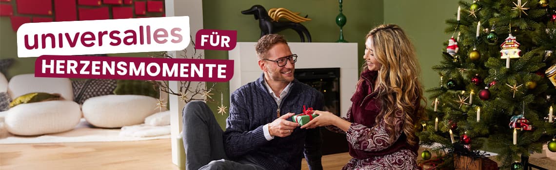 universalles für Herzensmomente – Paar sitzt vor Weihnachtsbaum und Kamin, Frau gibt Mann ein Geschenk
