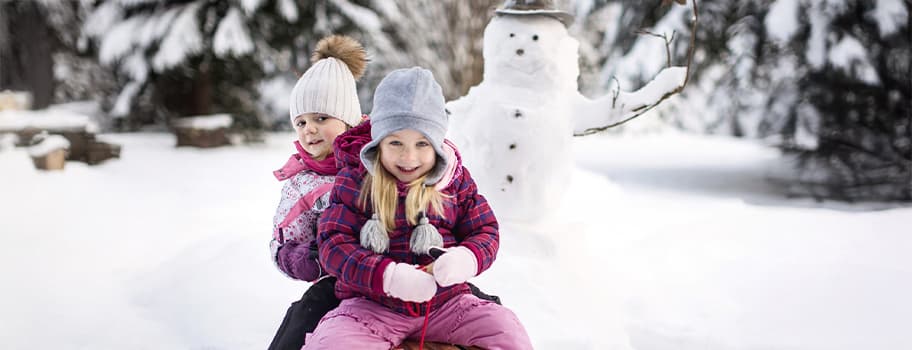 Zwei Kinder im Schnee spielen vor einem Schneemann.