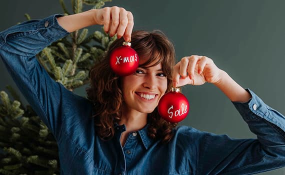 Person hält zwei rote Weihnachtskugeln vor Gesicht, eine mit "X" und eine mit "Sale" beschriftet, vor einem Weihnachtsbaum