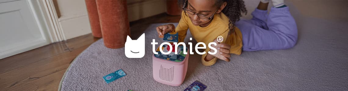 tonies – Kind in lila Hose und gelbem Oberteil spielt mit rosa Toniebox auf Teppich.