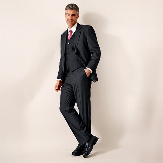 Eleganter Mann in schwarzem Anzug mit roter Krawatte und Weste, steht lässig an einer Wand.