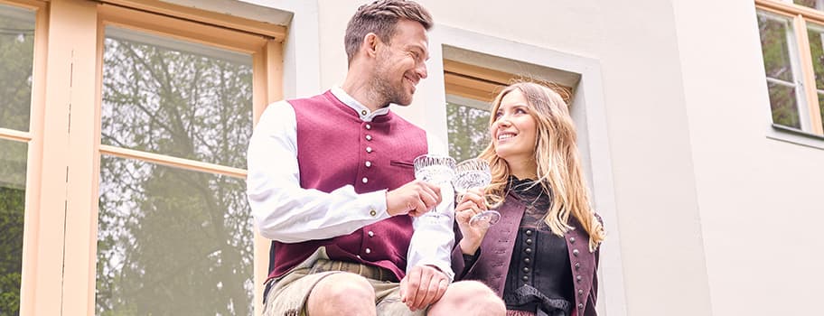 Paar in traditioneller Tracht stößt mit Gläsern an, sitzt vor einem Gebäude mit großen Fenstern.