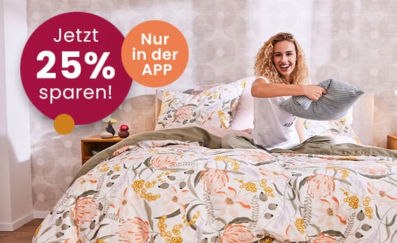Jetzt 25 % sparen! Nur in der APP – Frau sitzt auf einem Bett mit floralem Muster und hält ein graues Kissen.