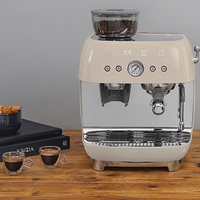 Cremefarbene Smeg-Kaffeemaschine auf Holztheke, daneben zwei Espressotassen und ein Buch.