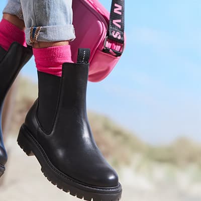 Person trägt schwarze Chelsea-Boots, pinke Socken und trägt eine pinke Tasche mit schwarzem Band.