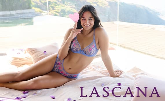 Frau in blauer und rosa Unterwäsche auf Bett mit Lascana-Logo, hält Herz vor Gesicht, Hintergrund mit Fenster und Natur.