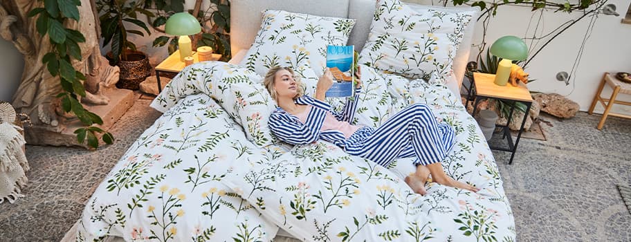 Person liegt in gestreiftem Pyjama auf floral gemustertem Bett, hält ein Buch. Pflanzen und Lampen im Hintergrund.