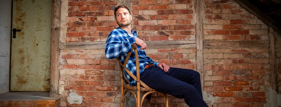 Mann in kariertem Hemd und dunkler Hose sitzt auf einem Stuhl vor einer Backsteinmauer