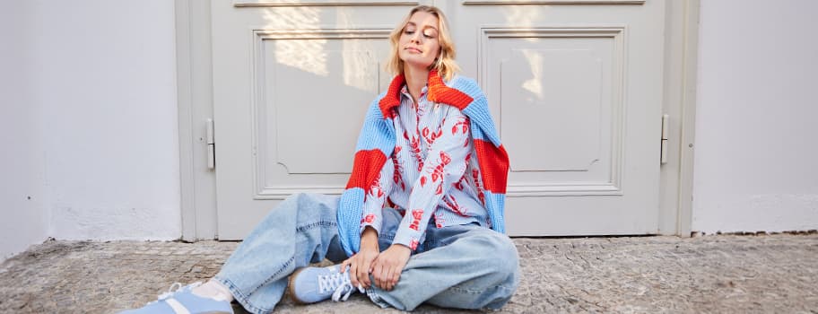 Person in farbenfrohem Hemd und blauer Jeans sitzt auf dem Boden vor einer Tür.