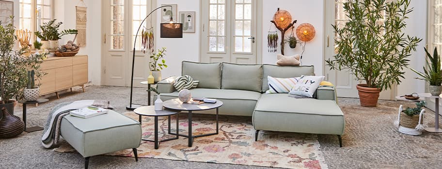 Modernes Wohnzimmer mit hellgrünem Sofa, Couchtischen, Pflanzen und Dekorationen vor weißen Türen und Fenstern.