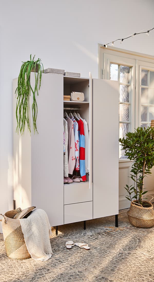 Offener Kleiderschrank mit bunten Kleidungsstücken, umgeben von Körben, Pflanzen und einem Fenster im Hintergrund.