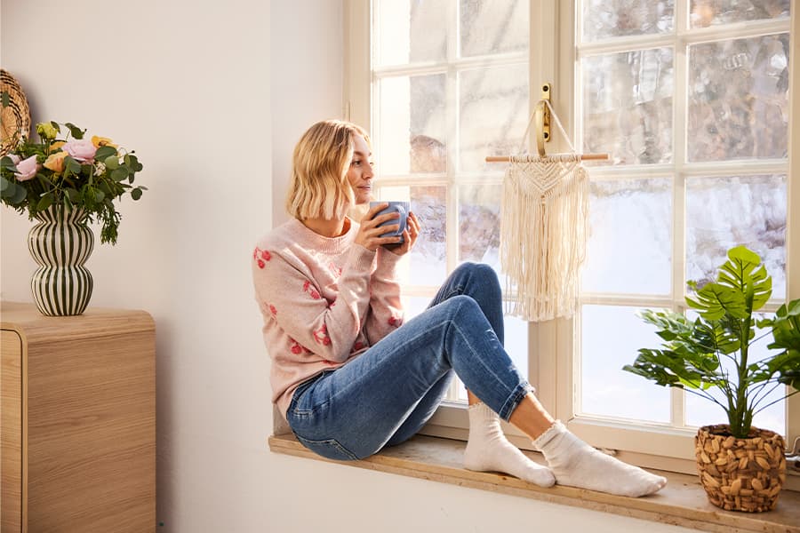 Frau sitzt am Fenster, hält eine Tasse, trägt einen rosa Pullover und blaue Jeans. Blumen und Pflanzen im Raum.