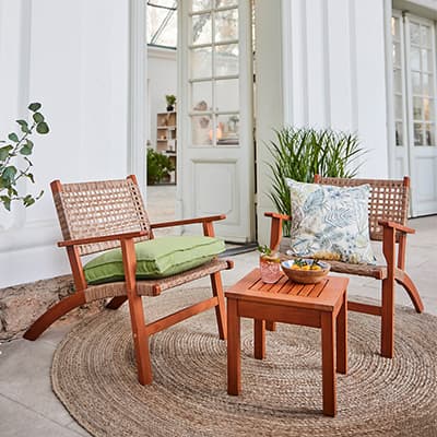 Zwei Holzstühle mit Kissen und ein kleiner Tisch auf einer Terrasse vor weißen Türen.
