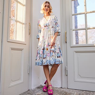 Frau in geblümtem Kleid und pinken Sandalen steht vor einer Tür, Gesicht unscharf.