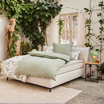 Hellgrünes Bett in hellem Raum mit Pflanzen und Statue, Fenster im Hintergrund.