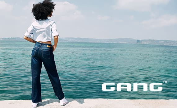 GANG – Person in Jeans steht mit dem Rücken zur Kamera am Ufer und blickt über das Wasser