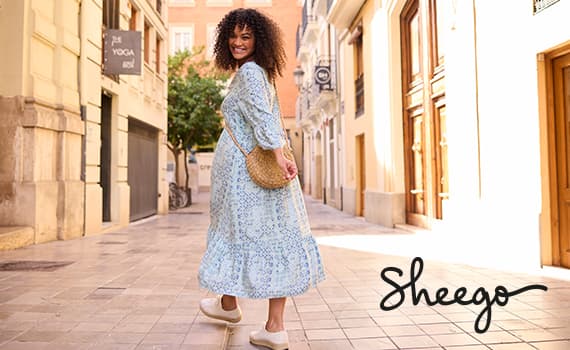 Sheego – Frau im hellen, gemusterten Kleid mit Umhängetasche geht durch eine sonnige Altstadtgasse