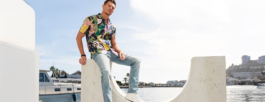 Mann in bunter Kurzarm-Hemd und Jeans sitzt auf weißer Skulptur am Wasser, im Hintergrund Boote und Gebäude.