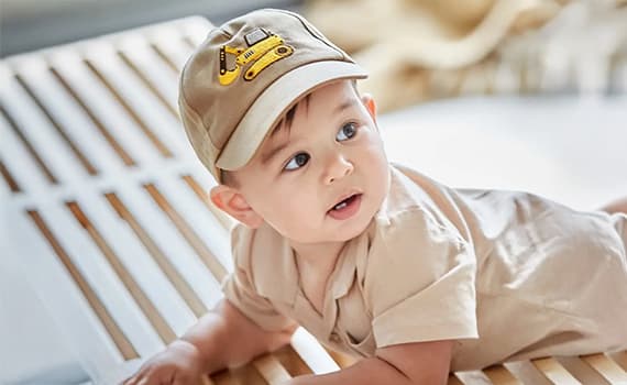 Baby in beigem Outfit und Mütze mit gelbem Bagger darauf liegt auf einer Holzbank.