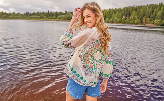 Frau steht am See und trägt eine gemusterte Bluse und Jeansshorts, sie schaut zur Seite.