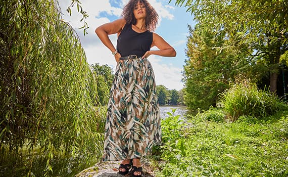 Frau in schwarzem Top und gemustertem Rock steht mit Händen an der Hüfte in einer grünen Landschaft.