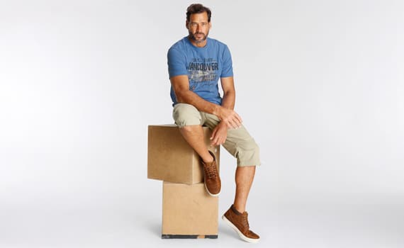 Mann in blauem Vancouver-T-Shirt und khakifarbenen Shorts sitzt auf zwei gestapelten Kartons.