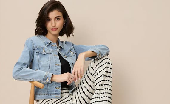 Frau in Jeansjacke und gestreifter Hose sitzt auf Stuhl vor beigefarbenem Hintergrund.