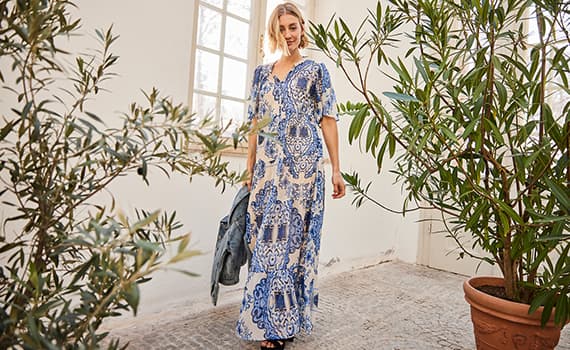 Frau in blau-weiß gemustertem Kleid steht in einem Raum mit Pflanzen und hält eine Jeansjacke in der Hand.