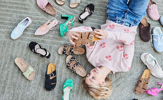 Frau liegt auf Teppich und umgeben von verschiedenen Schuhen, hält einen braunen Schuh in der Hand, trägt rosa Pullover und Jeans.