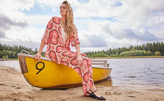 Frau in gemustertem Kleid sitzt auf gelbem Boot am Sandstrand vor See und Wald.