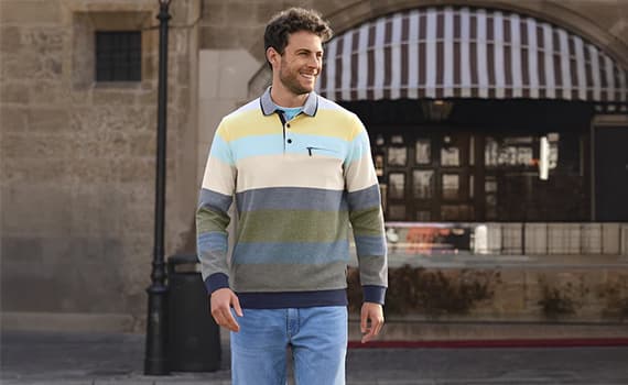 Mann trägt gestreiftes Langarmshirt und Jeans, geht vor Gebäude mit markanten Bogenfenstern.