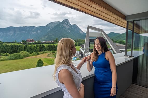 Zwei Frauen im Gespräch auf Balkon mit Blick auf grüne Landschaft und Berge im Hintergrund