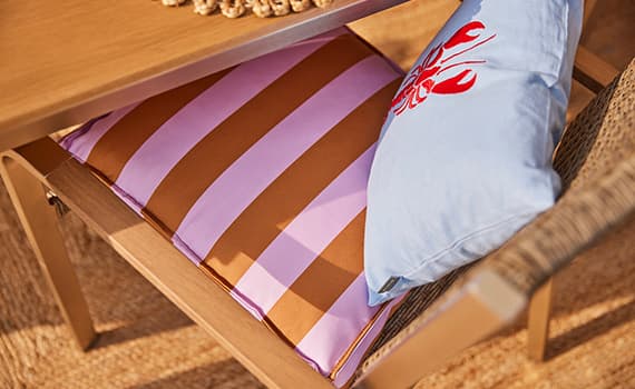 Gestreiftes Sitzkissen in Rosa und Braun auf Stuhl, daneben hellblaues Kissen mit rotem Hummermotiv unter Holztisch