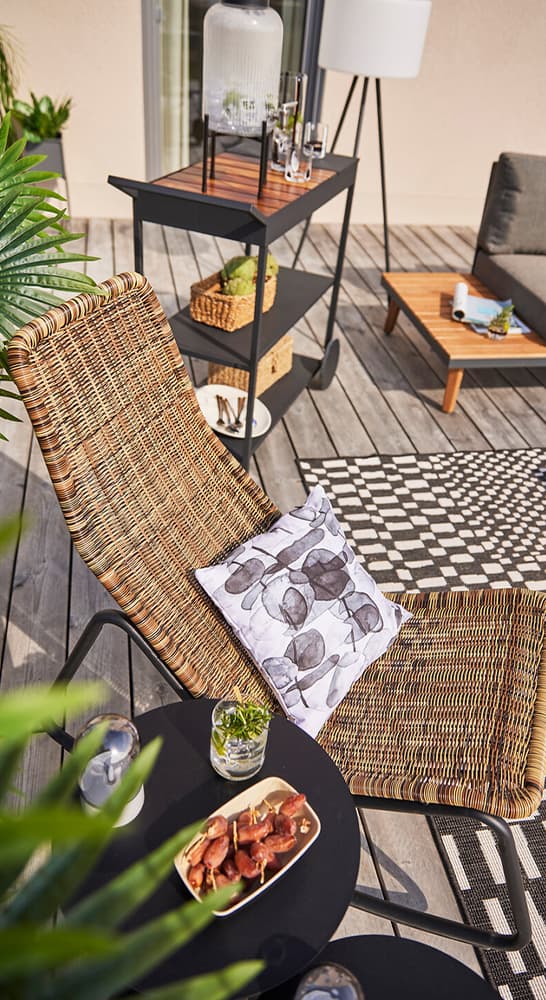 Liegestuhl mit gemustertem Kissen auf Terrasse, davor Beistelltisch mit Getränk und Snack, im Hintergrund Servierwagen