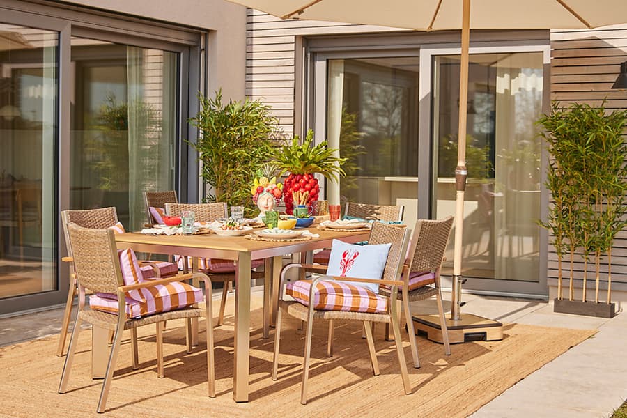 Gedeckter Gartentisch mit sechs Stühlen, buntem Geschirr, Obstschale und Blumen auf Terrasse mit Sonnenschirm
