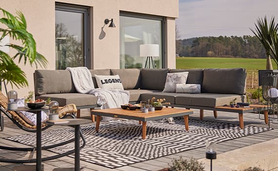 Ecksofa auf Terrasse vor Haus, Kissen mit Aufschrift „Legend“, Couchtisch auf gemustertem Teppich, Blick ins Grüne