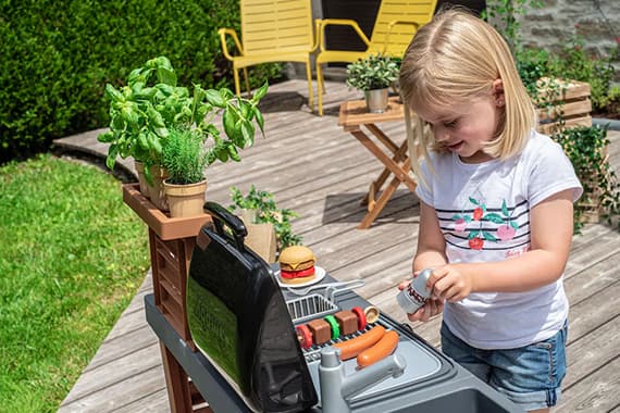 Kind spielt im Garten an einem Spielzeuggrill und würzt Würstchen, im Hintergrund Terrasse mit Stühlen und Pflanzen
