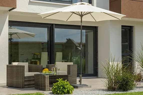 Terrasse mit Sitzgruppe, Tisch und Obstschale unter großem Sonnenschirm vor einem Haus mit bodentiefen Fenstern