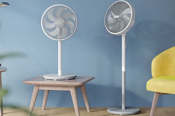 Zwei weiße Standventilatoren, einer auf einem niedrigen Tisch, einer auf dem Boden vor blauer Wand und gelbem Sessel